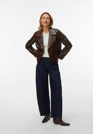 Femme debout les mains sur les hanches, portant une veste en cuir marron foncé, un haut blanc, un pantalon large bleu foncé, et des bottes marron foncé.
