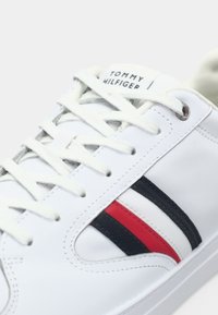 Adidași albi cu dungi roșii și bleumarin, design cu șireturi, suprafață sintetică netedă, logo pe limbă și talpă de cauciuc texturată.