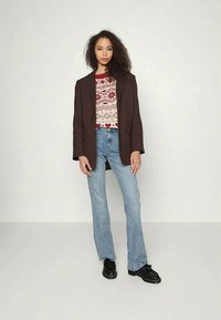 Blazer castanho ajustado sobre um suéter vermelho com padrão, jeans flare azuis e sapatos pretos. Inclui lapela em bico e bolsos.