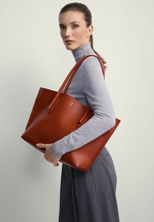 Kobieta w szarym golfie i spódnicach w prążki trzyma dużą, teksturowaną brązową torbę skórzaną na ramieniu.