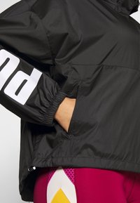 Svart lättviktig jacka med långa ärmar, elastiska muddars och framfickor; har djärv vit text på ärmen.