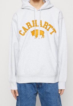 Persoon in een lichtgrijze hoodie met gele "CARHARTT" tekst en buffelafbeelding, gedragen met loszittende blauwe jeans.