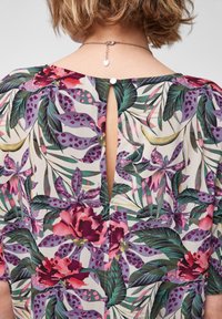Blusa floreale con apertura a goccia sul retro. Realizzata in tessuto leggero, con vivaci fiori viola e rosa e foglie verdi.
