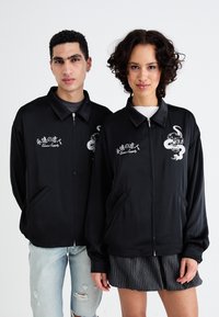 Črna zip-up jakna z gladko teksturo, ki ima spredaj belo grafiko in besedilo. Ima stranske žepe in ovratnik.