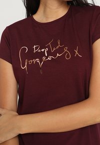 Burgundy t-paita lyhyillä hihoilla, jossa on kiiltävää metallista ruusukultaa oleva teksti "Drop Dead Gorgeous X" käsinkirjoitetussa tyylissä.