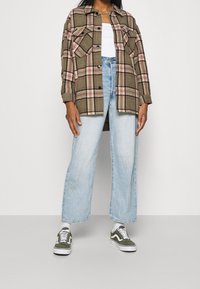 Camisa de franela a cuadros en verde y rosa, jeans de pierna ancha en azul claro y zapatillas verdes con acentos blancos. Dos bolsillos en el pecho de la camisa.