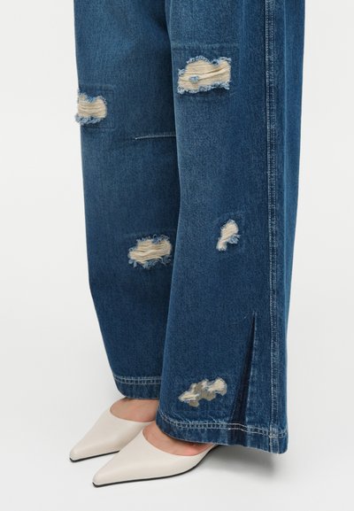 Jean bleu large usé avec des patchs effilochés et une fente latérale, porté avec des talons blancs pointus.