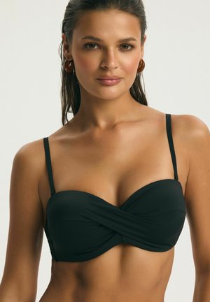 Frau mit nassem Haar, die ein schwarzes, verdrehtes Bandeau-Bikinioberteil und braune Creolen trägt, frontal vor einem einfarbigen Hintergrund.