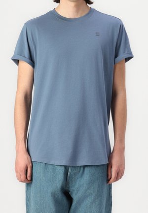 Person trägt ein schlichtes blaues T-Shirt mit kurzen Ärmeln und aufgekrempelten Ärmelbündchen sowie hellblaue Jeans vor weißem Hintergrund.
