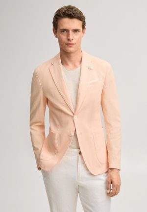 Młody mężczyzna stojący, ubrany w jasnoróżową marynarkę na beżową koszulę i białe spodnie, na jednolitym jasnym tle.