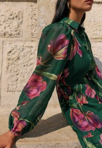 Žalia skaidri suknelė, puošta dideliais rožiniais ir violetiniais gėlių raštais, su ilgomis rankovėmis ir elastiniais manžetais, bei mygtukų užsegimu priekyje.