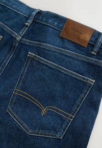 Tmavomodré džínsy z denimu s vreckom na zadnej strane, žltým prešívaním a hnedou koženou nášivkou s nápisom „DENIM SUPPLY“.