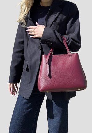 Persona che indossa un blazer a righe scure e jeans, che tiene in mano una borsa a tracolla texturizzata di colore rosso scuro con manici corti.