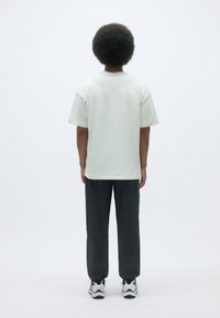 Light-colored short-sleeve t-shirt paired with dark, loose-fit pants and white sneakers, viewed from the back against a plain background.
