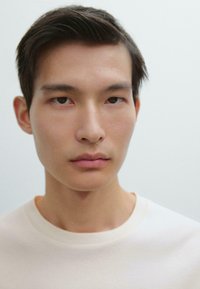 Primo piano di una persona con capelli corti e scuri, pelle liscia e espressione neutra, che indossa una t-shirt a girocollo di colore crema.