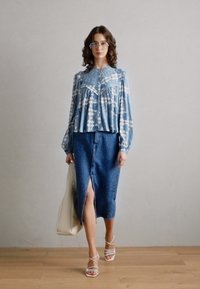 Lyseblå mønstret bluse med legg, kombinert med et jeansskjørt med høy splitt. Modellen har på seg sandaletter med stropper og bærer en beige bag.