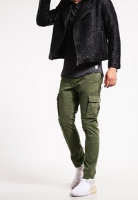 Czarna pikowana skórzana kurtka, dopasowana czarna koszula, oliwkowozielone spodnie typu cargo z kieszeniami oraz białe buty sportowe. Całość tworzy swobodny, miejski styl.