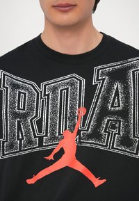 Μαύρο βαμβακερό μπλουζάκι με έντονο κείμενο "JORDAN" σε φθαρμένο λευκό, με μεγάλη κόκκινη σιλουέτα εν ός παίκτη μπάσκετ στον αέρα.