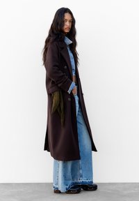 Brązowy długi płaszcz na dżinsowej kurtce, w połączeniu z szerokimi niebieskimi dżinsami. Czarne buty, zielony materiał w detalu w talii i teksturowane włosy.