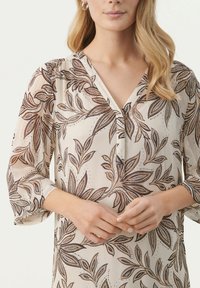 Blouse transparente en crème avec un motif de feuilles marron, décolleté en V, patte de boutonnage et manches trois-quarts. Tissu léger et aérien avec une texture douce.