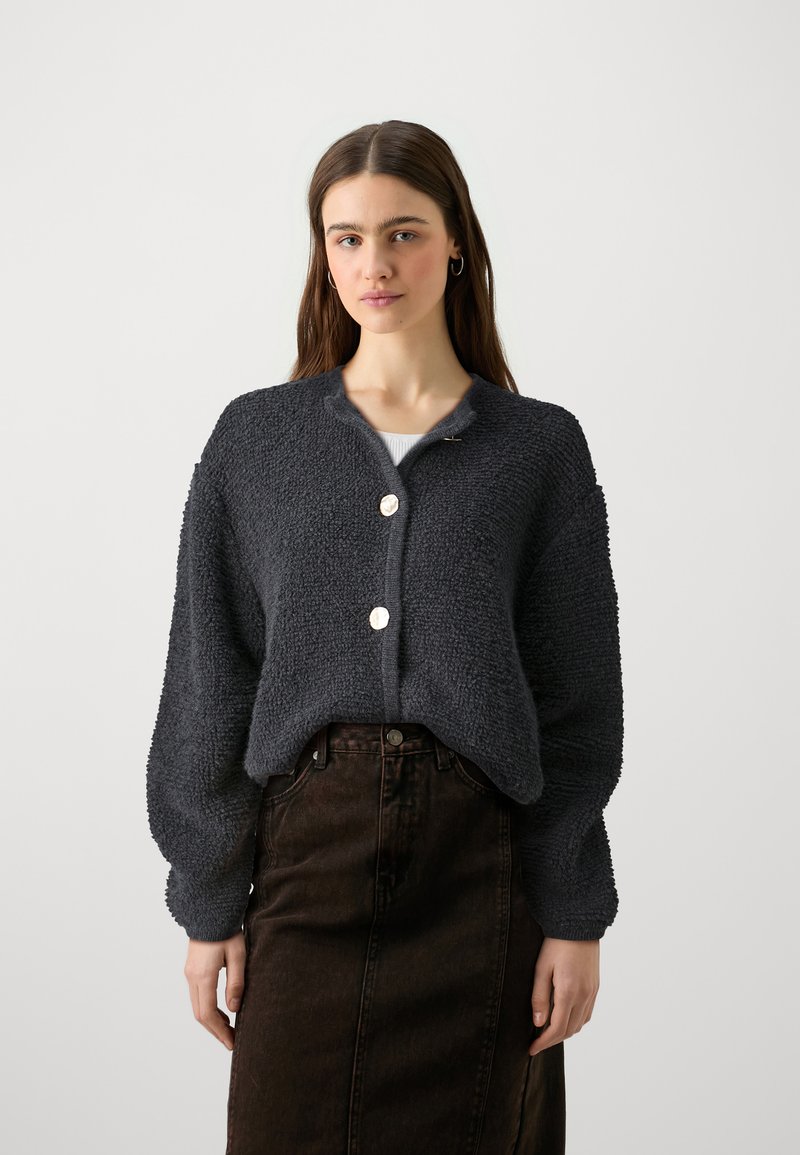 Σκούρος γκρίζος, υφασμάτινος κάρντιγκαν με στρογγυλή λαιμόκοψη και τρία μεγάλα κουμπιά. Χαλαρή εφαρμογή με χαμηλούς ώμους, φορεμένο πάνω από μια σκούρη φούστα.