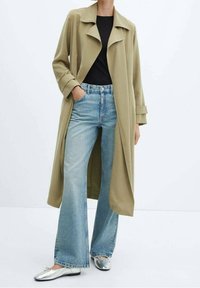 Trench coat di un leggero oliva con ampie revers, abbinato a un top aderente nero e jeans azzurri svasati. Ballerine argentate completano il look.