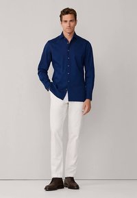 Camicia blu navy con bottoni realizzata in cotone, con maniche lunghe, collo con rever e bottoni marroni, abbinata a pantaloni bianchi e scarpe marroni.
