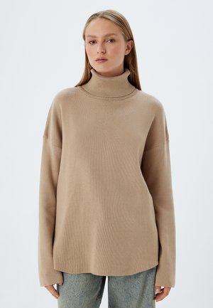 Pull à col roulé beige avec une coupe décontractée, col côtelé, texture douce et manches longues, associé à un jean bleu clair.