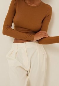 Kvinde iført en tætsiddende ribbet brun langærmet crop top og høj talje løse cremefarvede bukser, der poserer med den ene arm over kroppen.