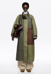 Abrigo largo a bloques de color en verde oliva y marrón con un cuello alto, bolsillos laterales y un logo. Combinado con pantalones de pierna ancha beige y zapatos marrones.