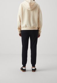 Persona con una sudadera beige clara, pantalones jogger negros y zapatillas negras de pie de espaldas a la cámara sobre un fondo limpio.