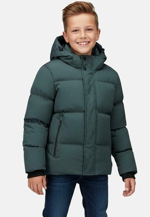 Jongen glimlachend, draagt een groen gewatteerd winterjack met kap en blauwe spijkerbroek tegen een effen achtergrond.