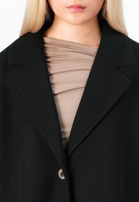 Woman with long blonde hair wearing a black coat over a taupe ruched top, shown from neck to mid-torso against a white background.