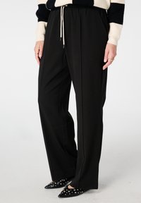 Pantalon noir à jambes larges avec taille élastique et cordon de serrage. Tissu lisse, poches latérales, accompagné de chaussures noires pointues ornées de clous dorés.
