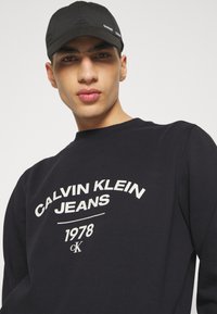 Cappello nero con dettagli del logo, indossato con una felpa blu marina con testo bianco: "CALVIN KLEIN JEANS 1978" in un font audace.