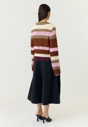 Femme aux longs cheveux foncés portant un pull en maille rayé marron, rose et crème, un pantalon culotte noir à jambes larges et des mules noires à talons, debout devant un fond blanc.