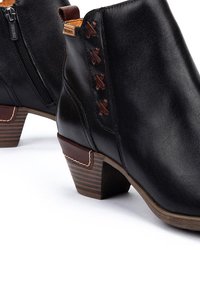 Botas de piel negras hasta el tobillo con tacones apilados marrones, cremallera lateral y un detalle decorativo de puntadas cruzadas marrones en el lado exterior.