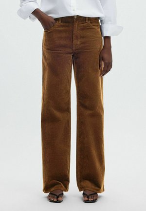 Personne portant un pantalon large en velours côtelé marron, une chemise blanche à manches longues avec les poignets retroussés, et des sandales noires à bouts ouverts, main dans la poche.