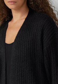 Czarny sweter z dzianiny z textured wzorem, zapinany na przód, w luźnym fasonie, noszony na czarnej bluzce. Miękka, przytulna tkanina.