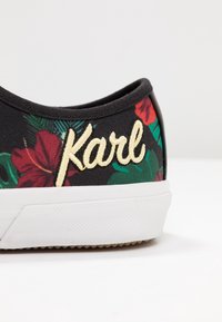 Mustat tennarit, joissa on kukkakuosi ja keltaisella kirjailtuna "Karl" valkoisen teksturoidun pohjan päällä valkoista taustaa vasten.