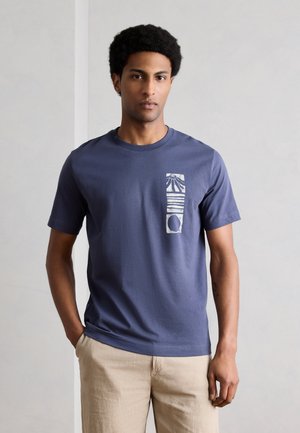 Άνδρας με σκούρο μπλε T-shirt με κάθετα λευκά γραφικά στο αριστερό στήθος και μπεζ παντελόνι, στέκεται με το ένα χέρι στην τσέπη.