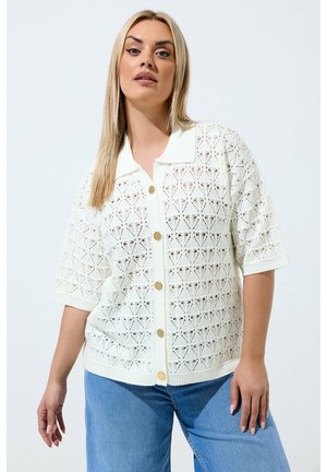 Kvinde iført en hvid kortærmet hæklede skjorte med knapper og krave, kombineret med blå jeans, stående mod en ensfarvet baggrund.