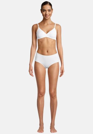 Donna con capelli raccolti che sorride, indossa un reggiseno bianco e mutandine a vita alta, in piedi a piedi nudi contro uno sfondo semplice.