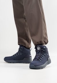 Marine hiking boots met een gestructureerde afwerking, metalen veterogen en reflecterende veters, gecombineerd met bruine elastische cuffbroeken.