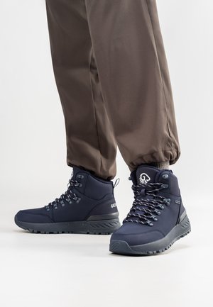 Marinefarbene Wanderstiefel mit strukturierter Oberfläche, metallischen Ösen und reflektierenden Schnürsenkeln, kombiniert mit braunen, elastischen Hosen mit Bündchen.