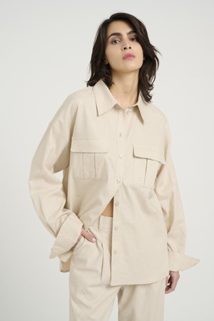 Camicia beige oversize con bottoni realizzata in un tessuto texturizzato, caratterizzata da due tasche frontali e ampi polsini, abbinata a pantaloni coordinati.