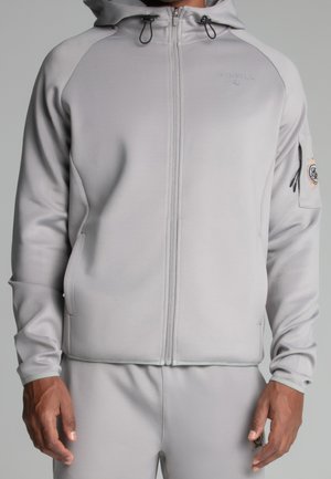 Mann trägt hellgrauen Reißverschluss-Hoodie mit Kordeln und passende Jogginghose, steht vor einfachem Hintergrund.