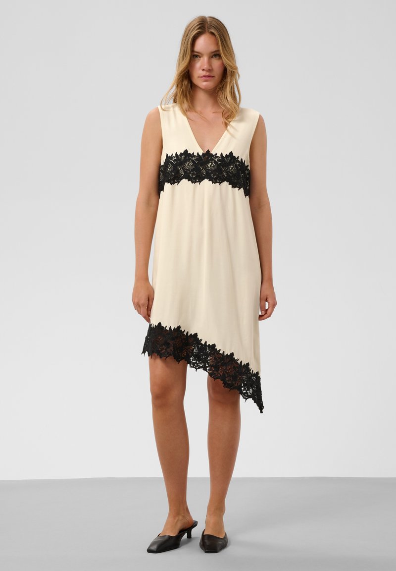Femme portant une robe crème sans manches avec bordure en dentelle noire sur la poitrine et ourlet asymétrique, associée à des chaussures noires à talons bas.