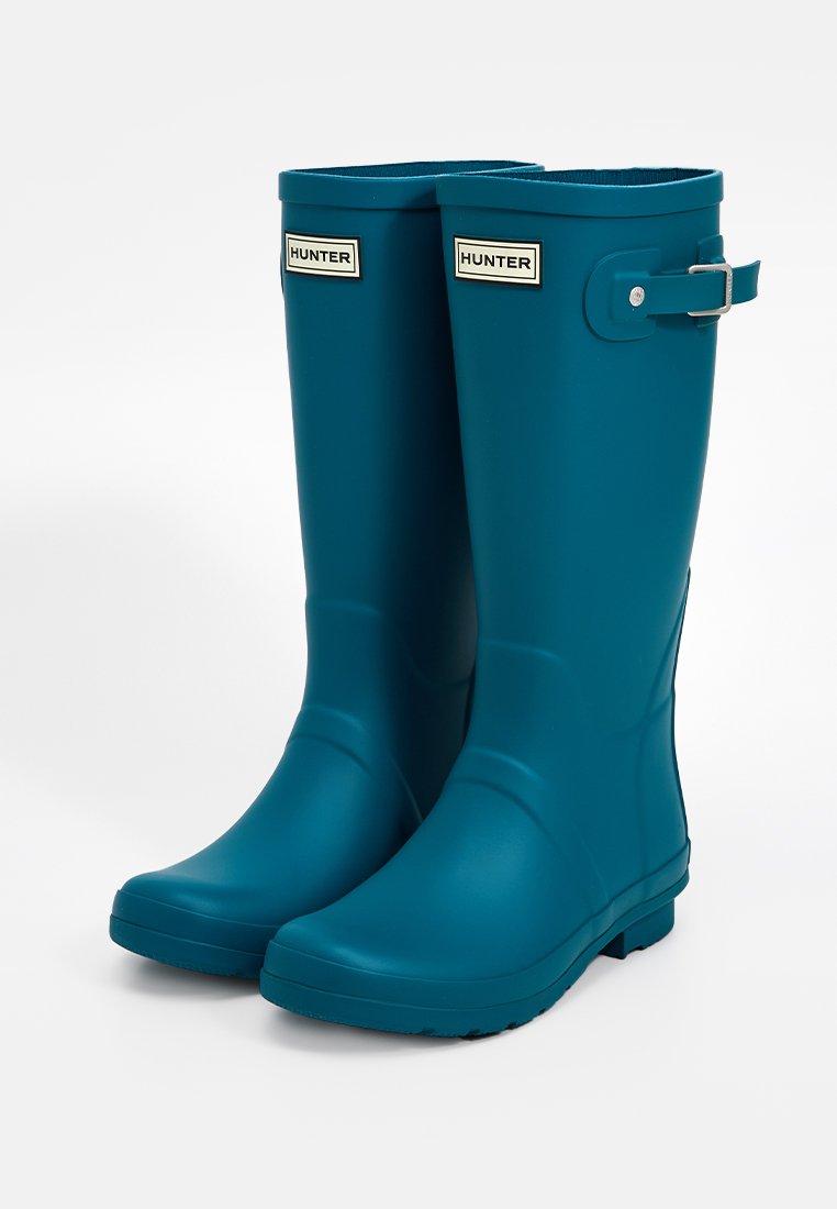 Botas de lluvia de goma en color verde azulado con acabado mate, puntera redondeada, correa con hebilla de metal y etiqueta con el logotipo cerca de la parte superior.