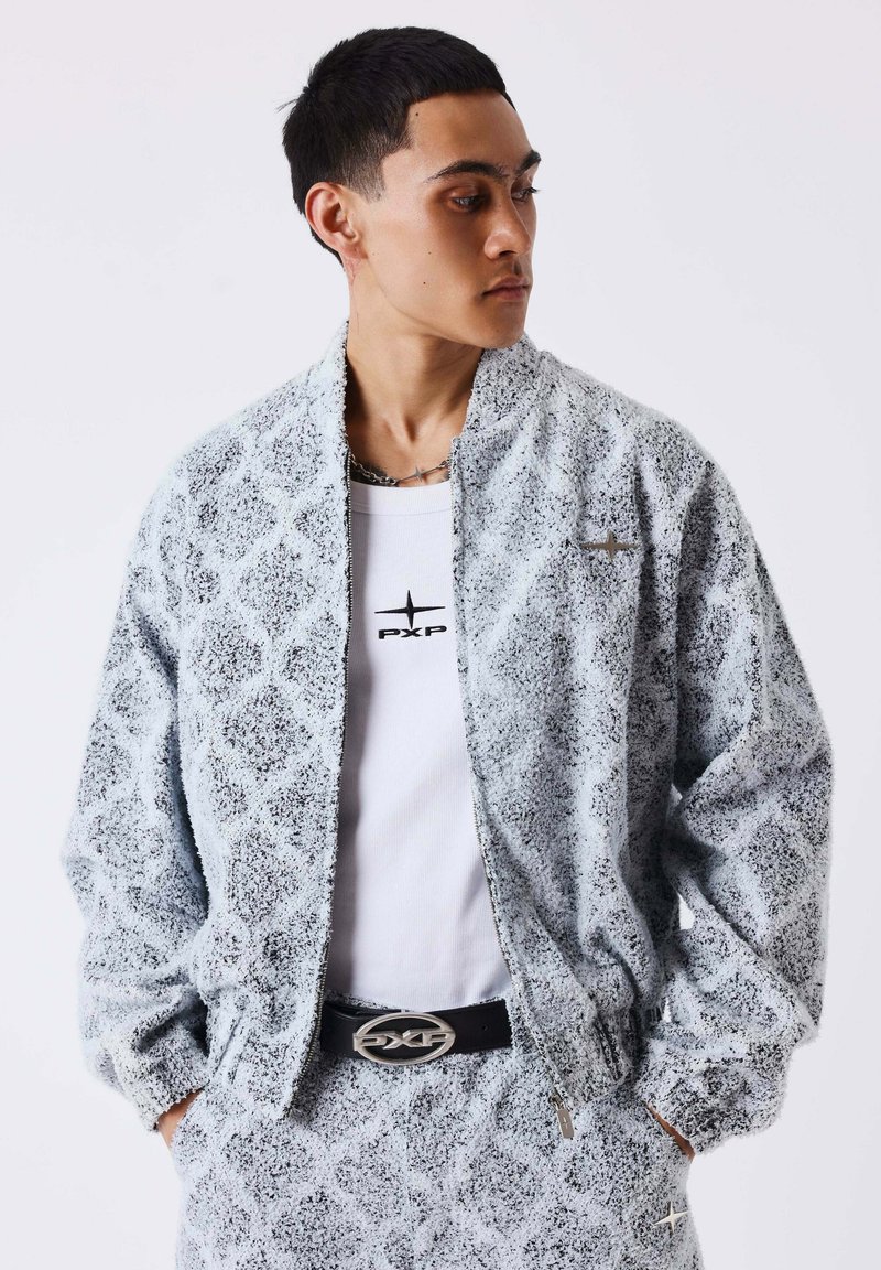 Jeune homme portant une veste et un pantalon assortis gris clair texturés avec des motifs géométriques, t-shirt blanc, ceinture noire, regardant vers sa gauche.